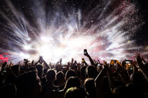 Excited crowd with raised hands and phones capturing the moment at a vibrant night music festival, illuminated by bright stage lights, confetti, and fireworks.